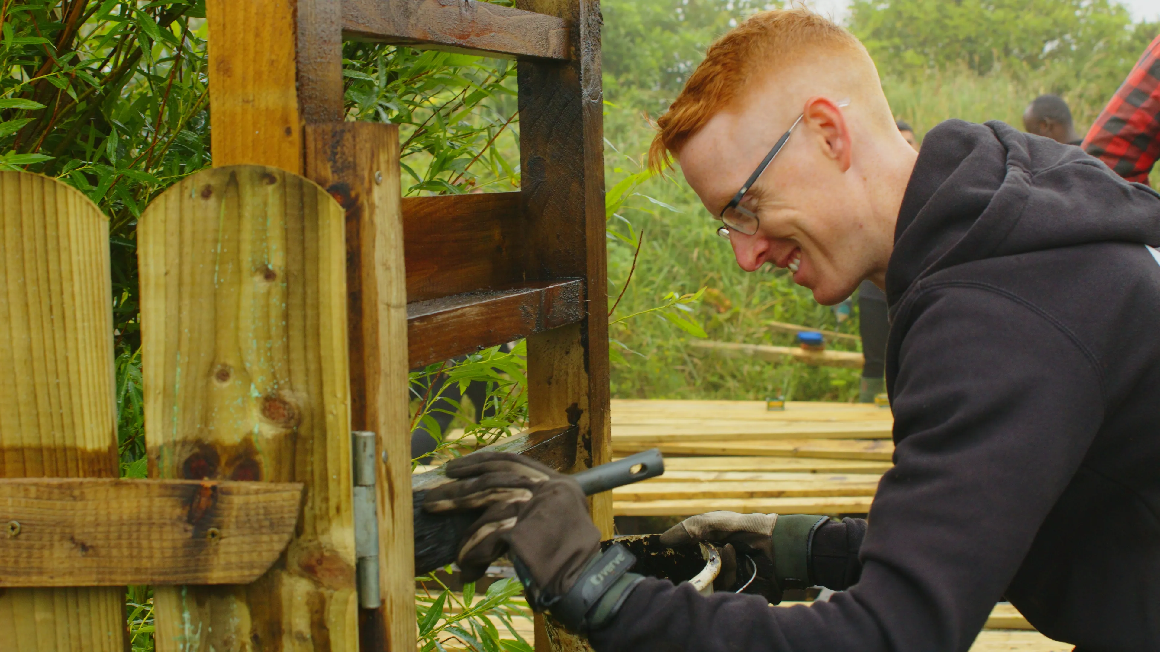 A person wearing glasses and gloves enthusiastically paints a wooden fence outdoors, surrounded by greenery, conveying a sense of focus and satisfaction.